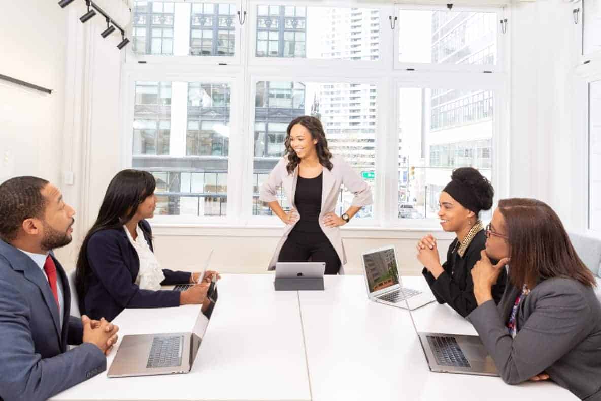 Traits and behaviours of successful leaders. Woman standing at the head of conference table addressing others seated.